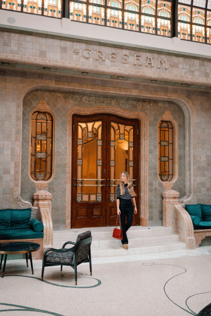 Olga stands on the marble steps beneath the patterned glass and stonework of the Gresham Palace lobby.