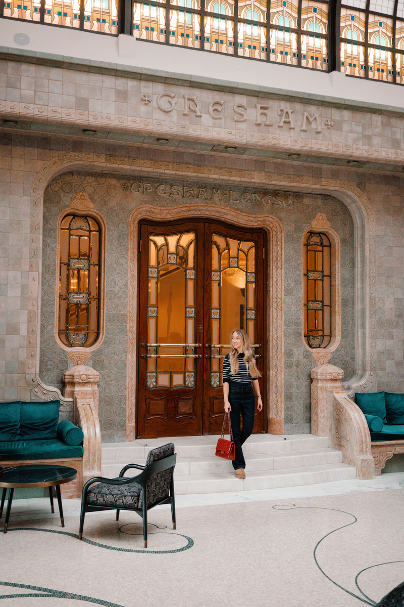 Olga stands on the marble steps beneath the patterned glass and stonework of the Gresham Palace lobby.