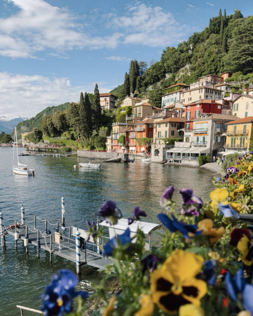 Colorful waterfront houses in Varenna beside Lake Como, with flowers and a small sailboat in the foreground.