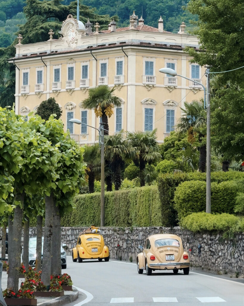 Two vintage Volkswagen Beetles drive past a pale lakeside villa surrounded by palms and clipped hedges.