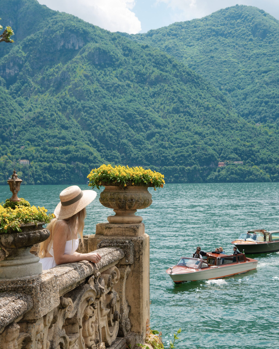 Olga, wearing a white dress and straw hat, looks across Lake Como from a stone terrace at Villa del Balbianello.