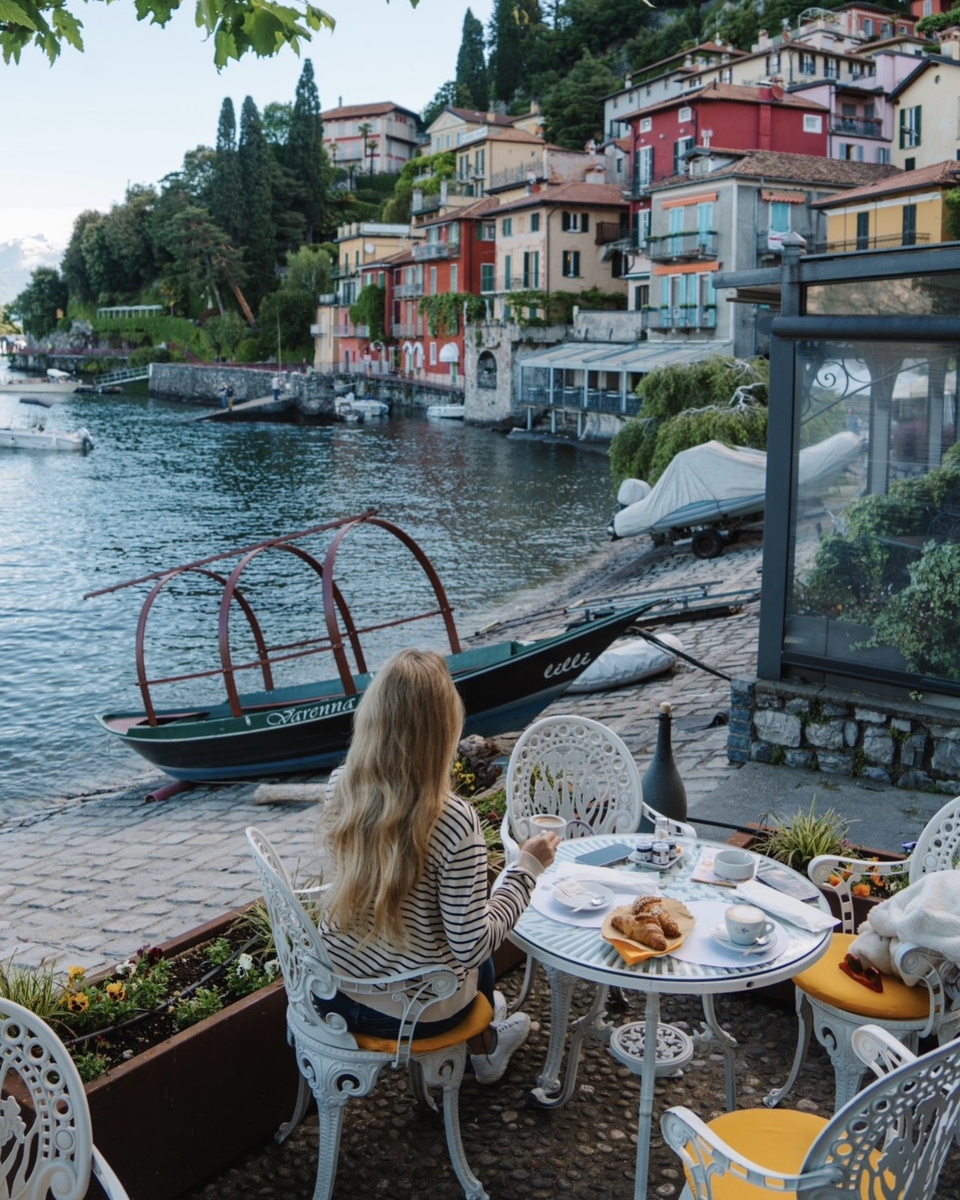 Olga has breakfast at a waterside table in Varenna, with colorful houses and a traditional boat behind her.
