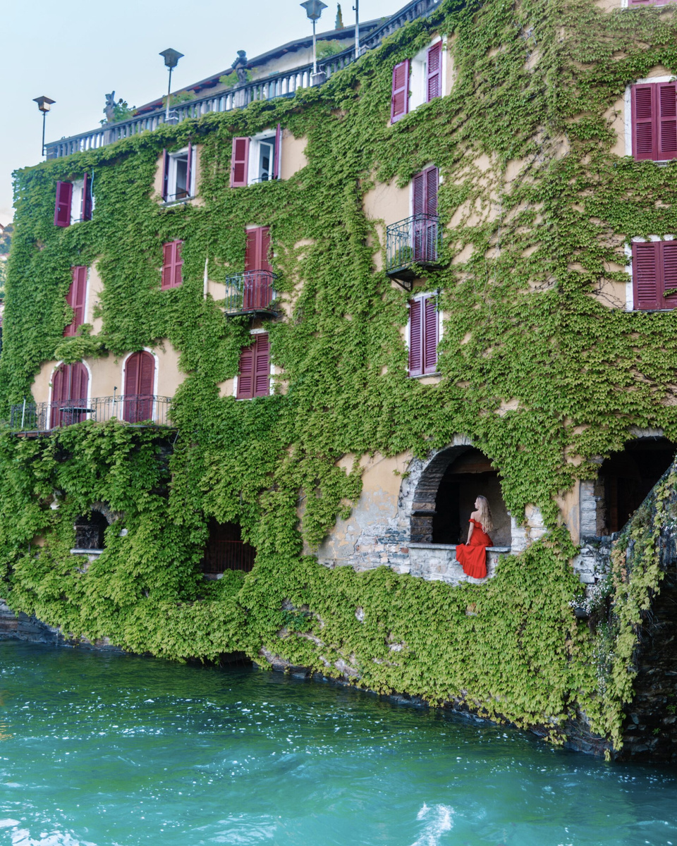 Olga, wearing a red dress, stands in an ivy-covered waterside arch in Nesso.
