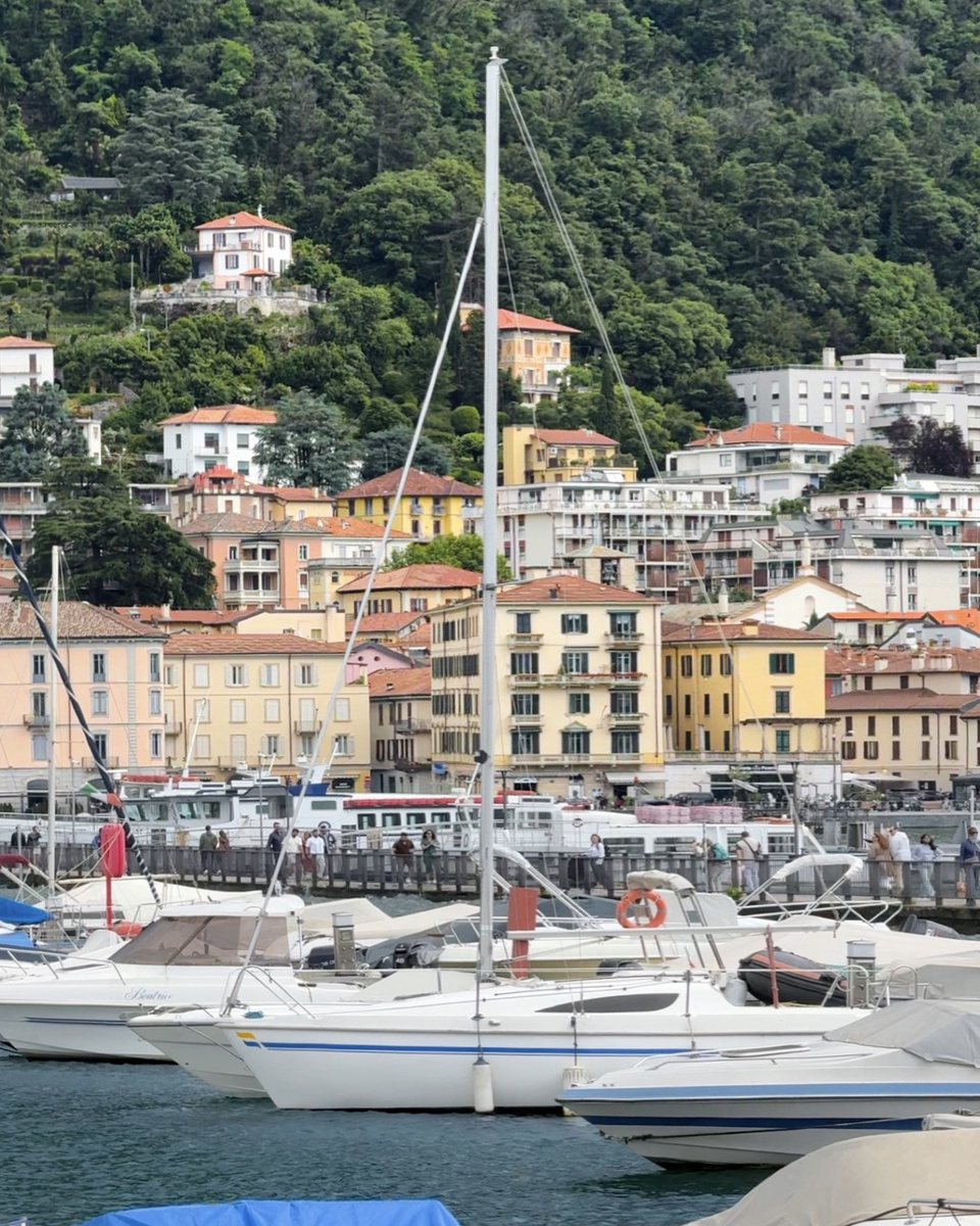 Sailboats in Como's marina, with colorful buildings climbing the wooded hillside behind them.