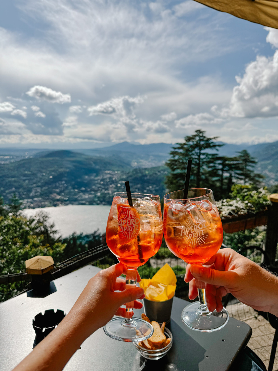Two glasses of Aperol Spritz are raised above Lake Como from a hillside terrace in Brunate.