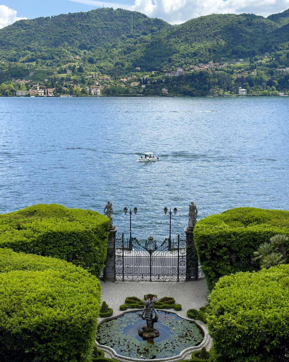 A formal lakeside garden with clipped hedges, a fountain, statues, and wrought-iron gates overlooking a passing boat.