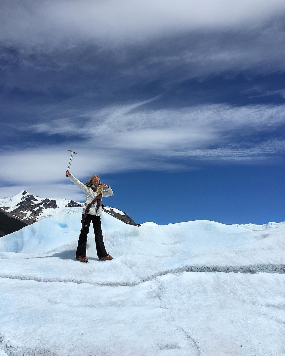 Olga raising a trekking pole on the Perito Moreno Glacier beneath dark mountains.
