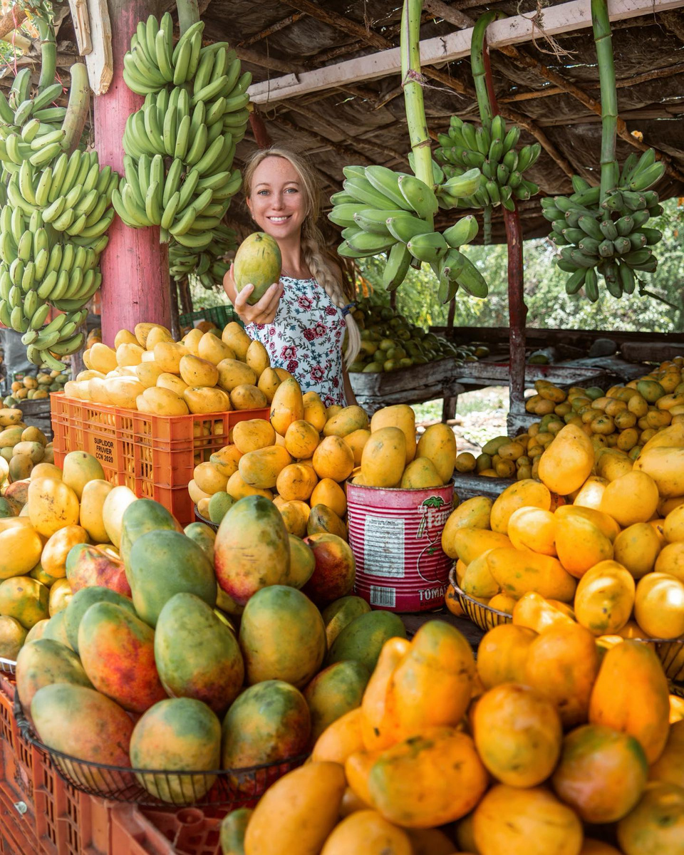 Olga holding a ripe mango at a roadside fruit stall in Baní.