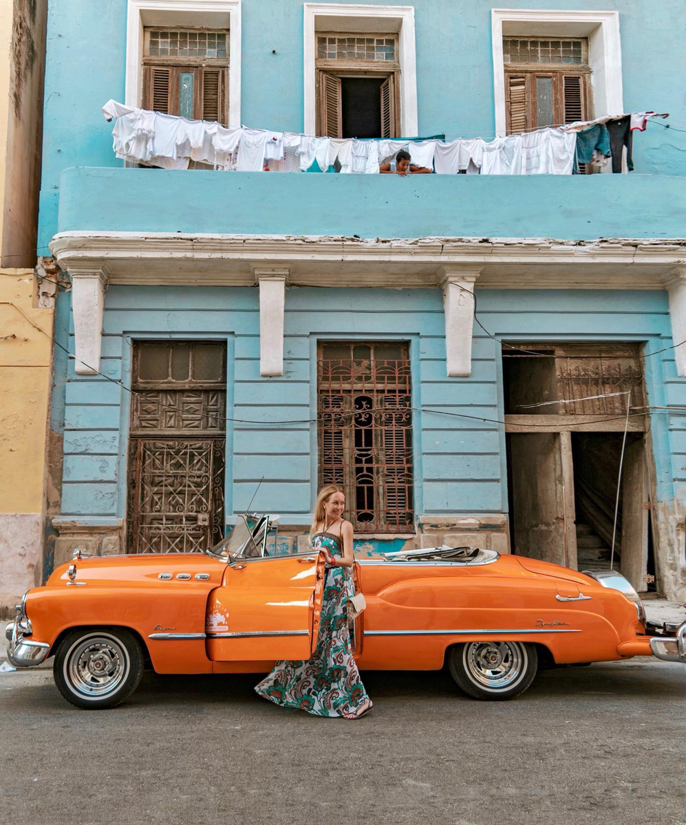 Olga in a patterned dress beside an orange vintage car and blue Havana townhouse.