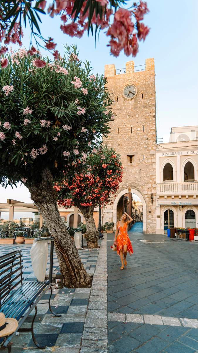Olga crossing a sunny square beneath flowering trees in Taormina.