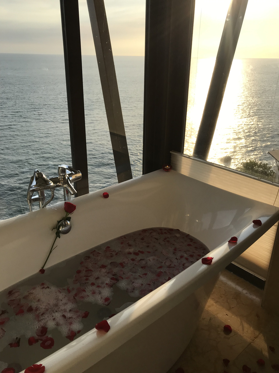 A bathtub filled with rose petals stands against tall windows overlooking the sunlit Pacific.