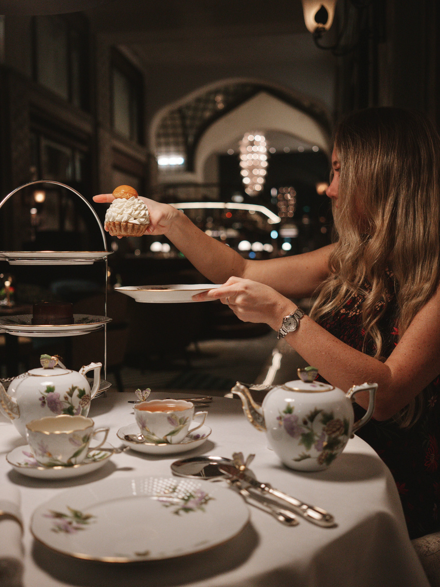 Olga, wearing a red-and-black evening dress, lifts a pastry from the afternoon-tea stand at MÚZSA.