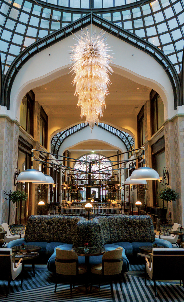 The soaring glass-roofed lobby of Four Seasons Gresham Palace, with a sculptural chandelier above blue seating and the MÚZSA bar.