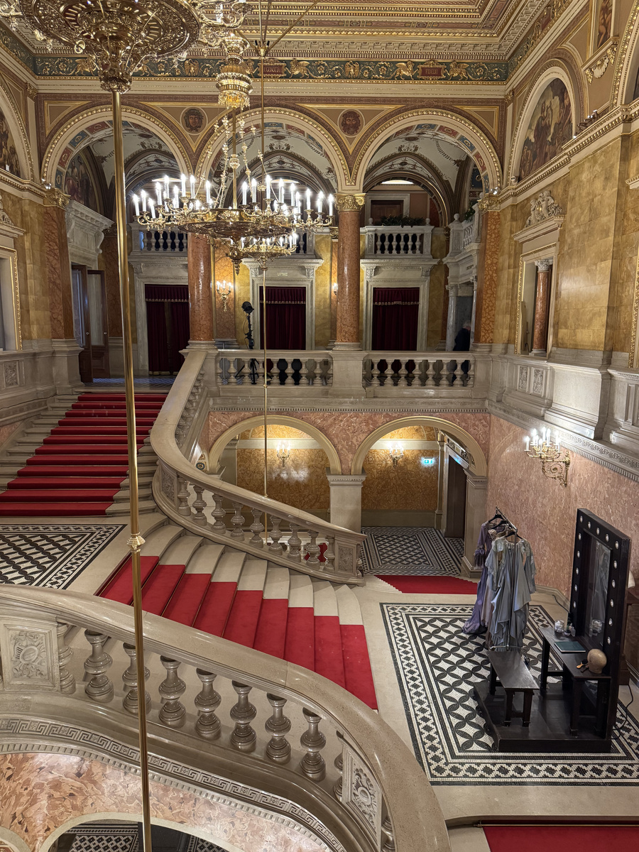 A red-carpeted marble staircase curves through the richly painted interior of the Hungarian State Opera, with costumes waiting beside a mirror.