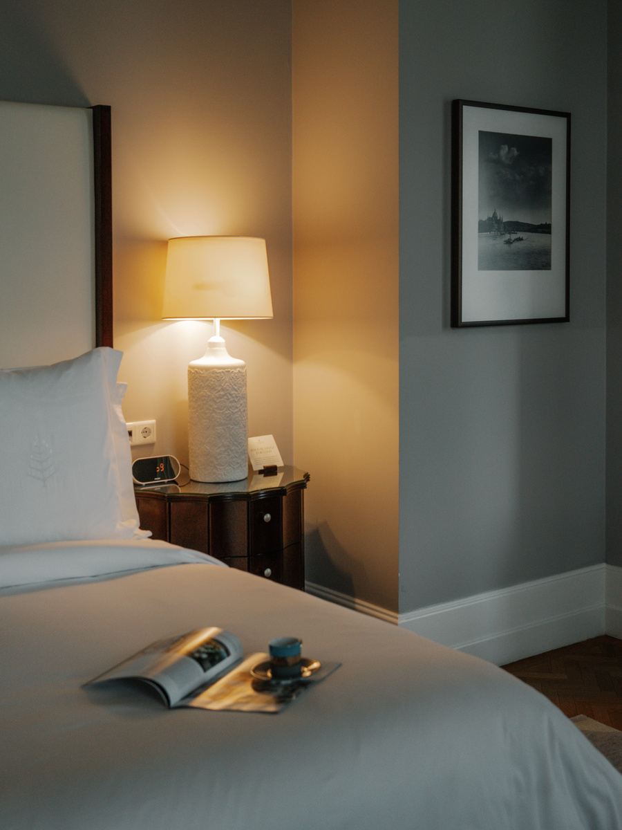 A pale guest-room bed is lit by a bedside lamp, with a book and chocolates resting on the cover.