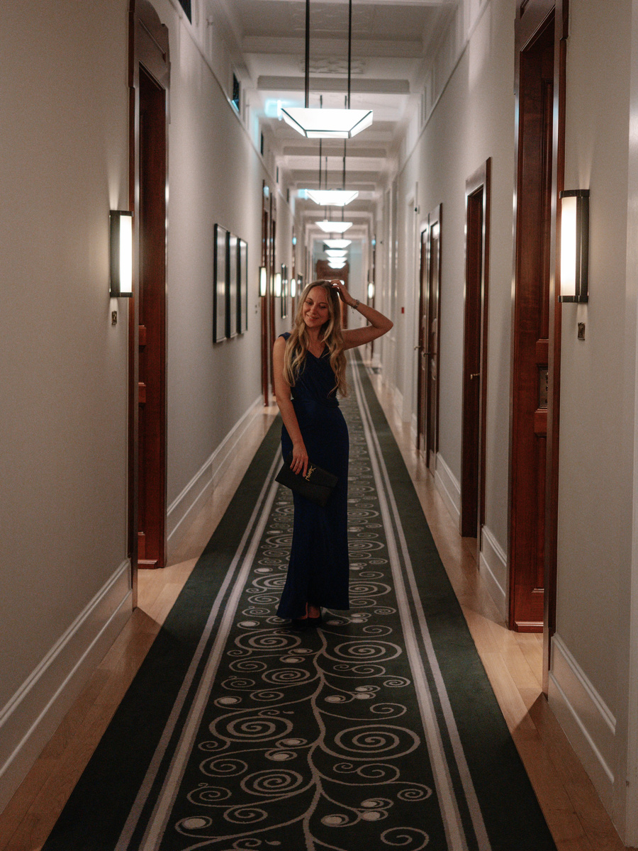 Olga wears a dark blue evening gown in a long Four Seasons Gresham Palace corridor, with pendant lights receding behind her.