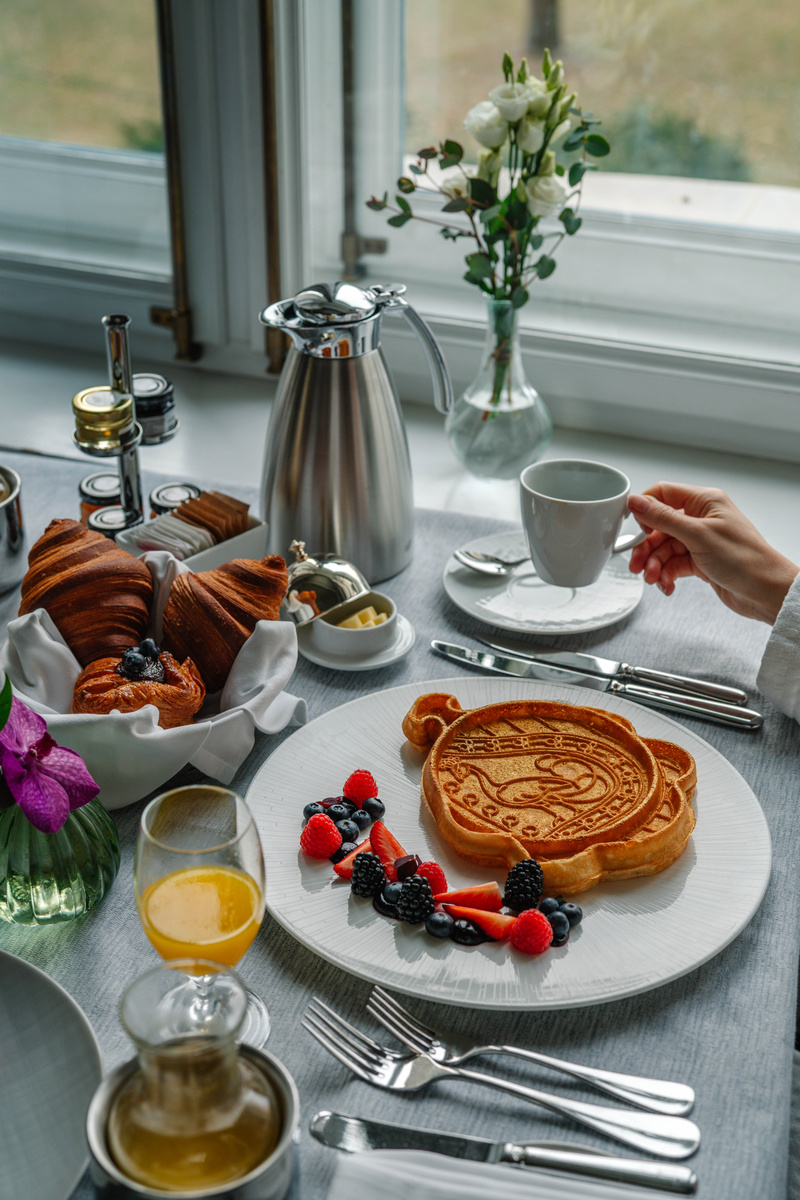 A breakfast waffle stamped with the Gresham Palace peacock is served with berries beside coffee, pastries and flowers.