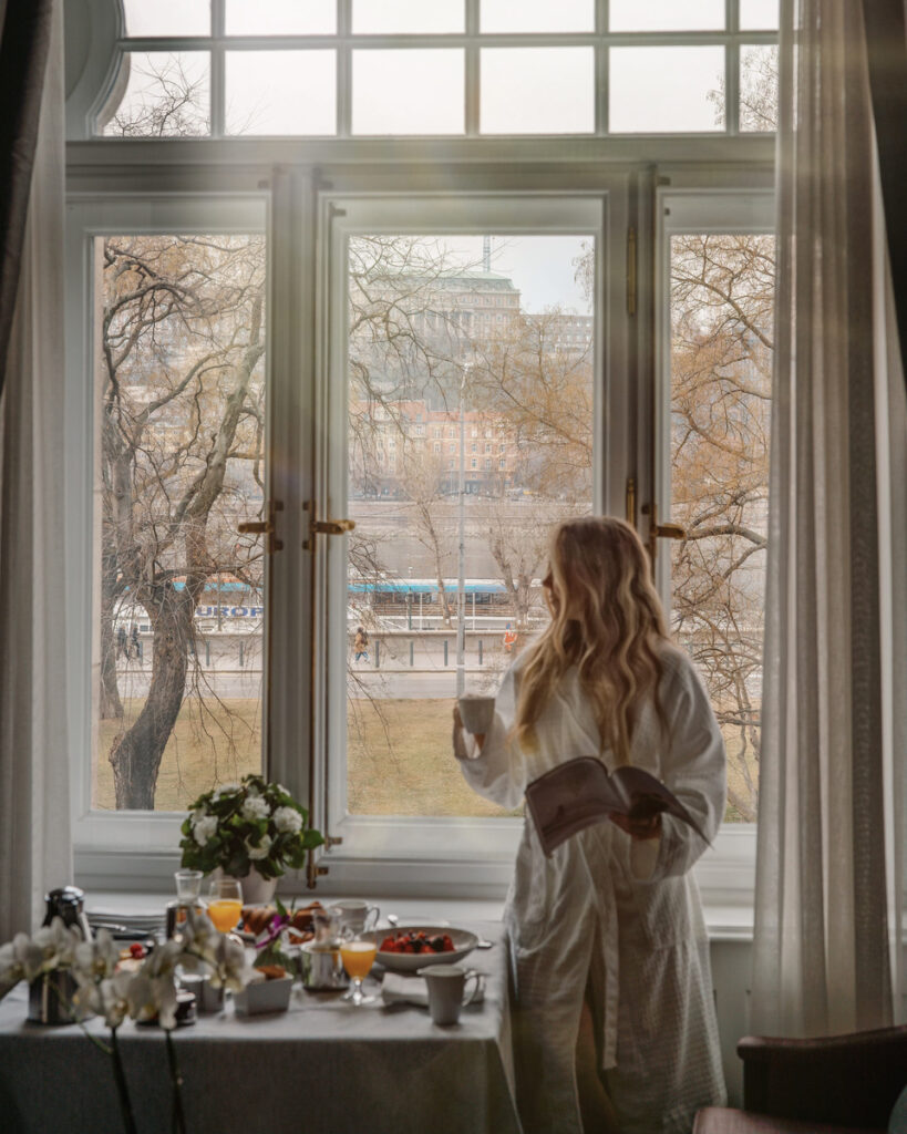 Olga stands with coffee beside an in-room breakfast table and tall windows overlooking Buda.