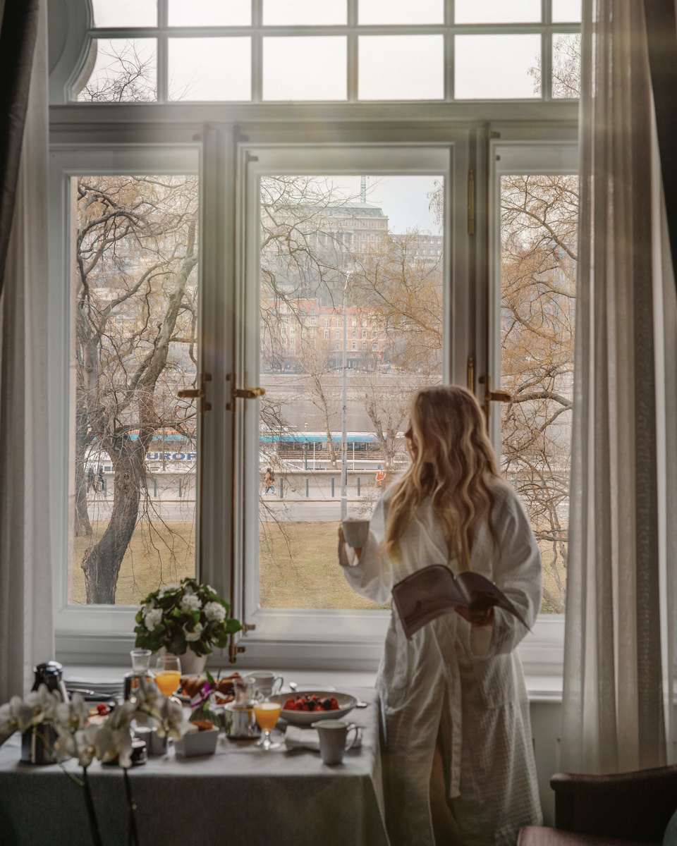 Olga stands with coffee beside an in-room breakfast table and tall windows overlooking Buda.