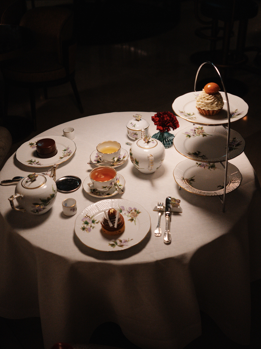 An afternoon tea table is laid with porcelain, cakes, sandwiches, scones and flowers beneath a pool of warm light.