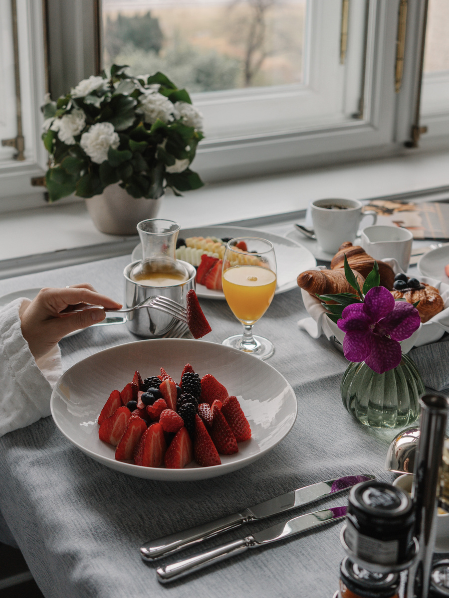 A bowl of strawberries and a glass of orange juice sit beside coffee, croissants and white flowers.