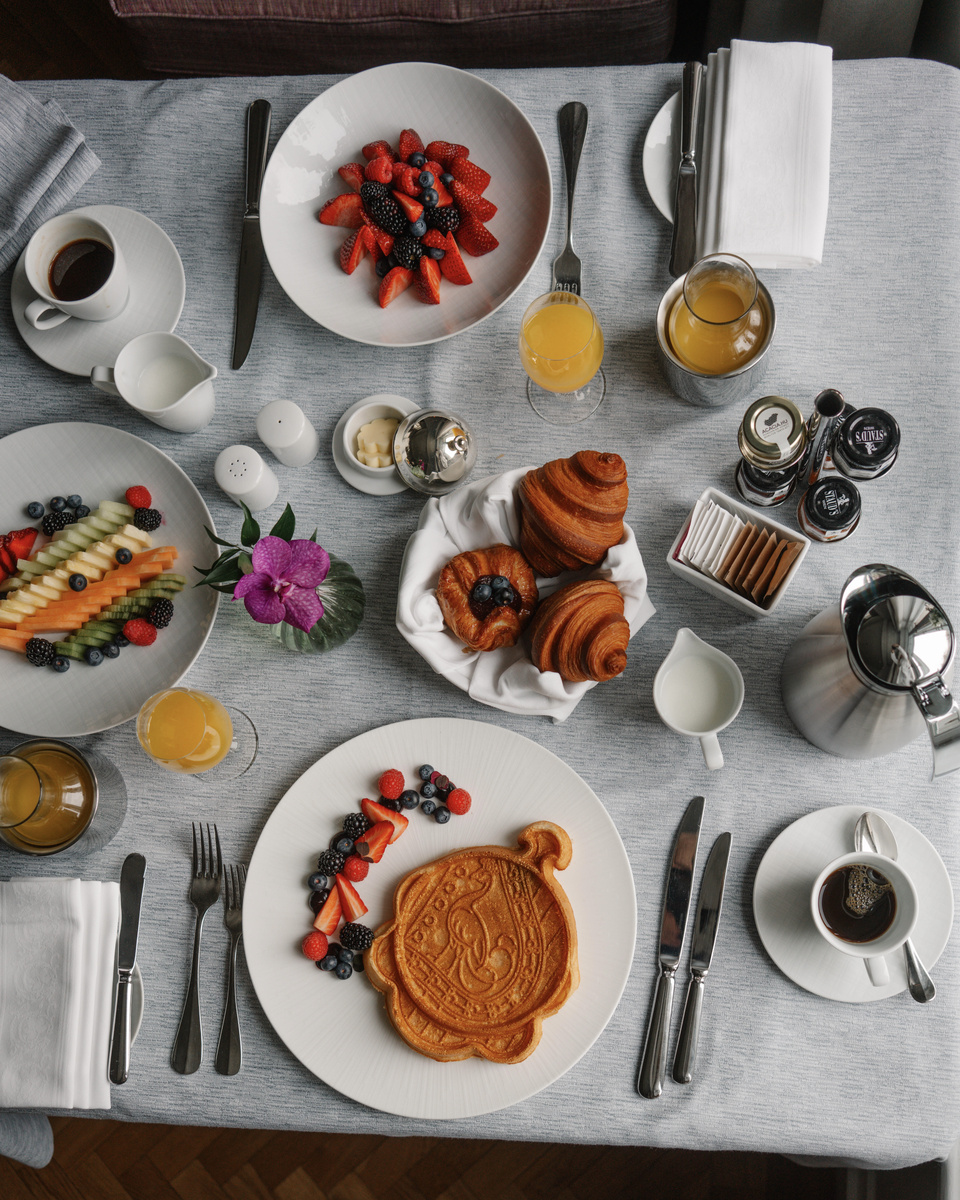 An overhead breakfast table is arranged with waffles, fruit, pastries, coffee, eggs, juice and yoghurt.