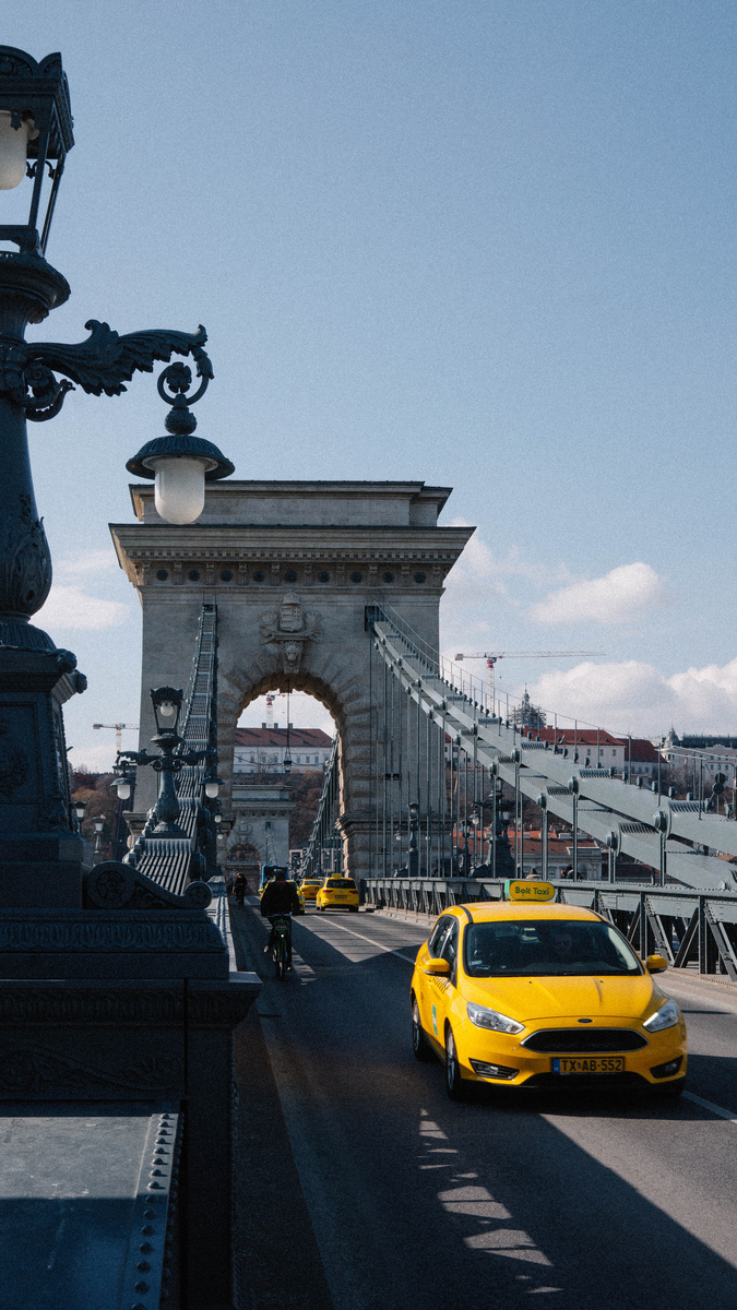 The stone entrance of Budapest’s Chain Bridge rises against a blue sky, with yellow taxis passing below.