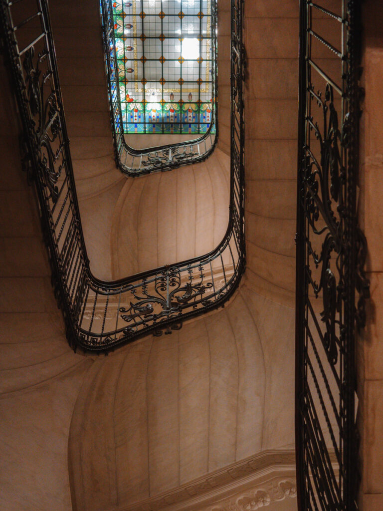 Curving wrought-iron stair rails rise towards a floral stained-glass ceiling at Four Seasons Gresham Palace.