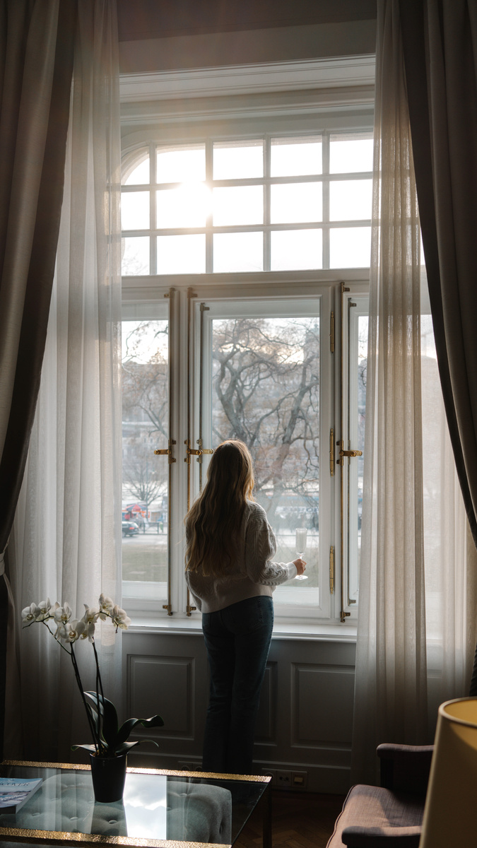 Olga stands at a tall hotel window with a drink as winter sun enters between sheer curtains.