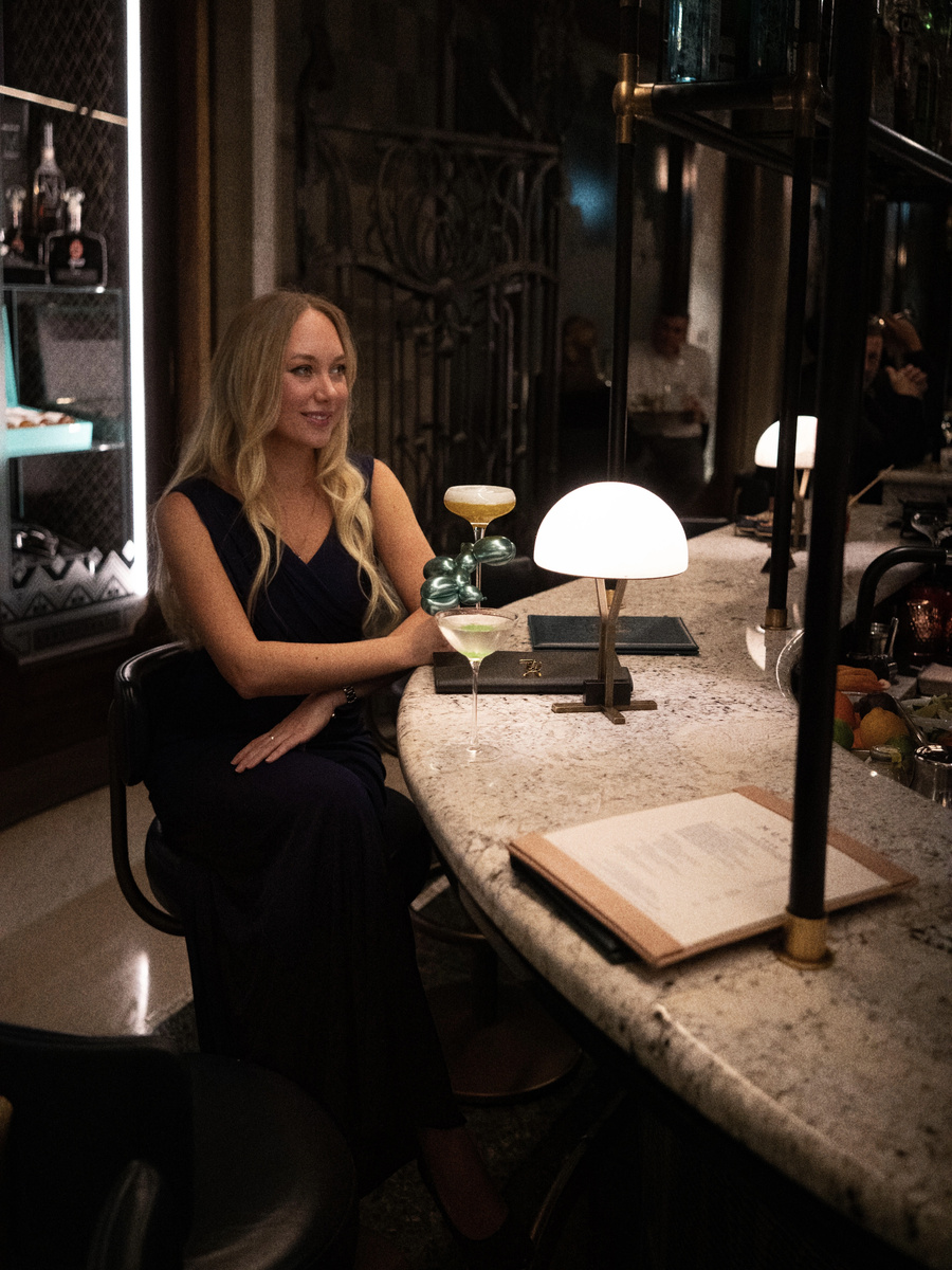 Olga sits at the curved MÚZSA bar beside two sculptural cocktails and a white table lamp.