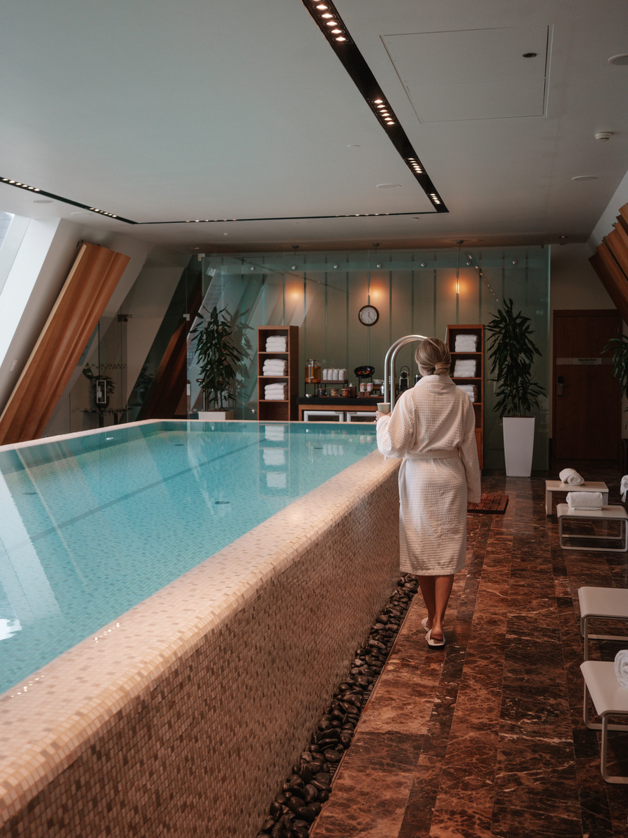 Olga walks beside the indoor pool in a white robe, with the water and sloping glass windows ahead.