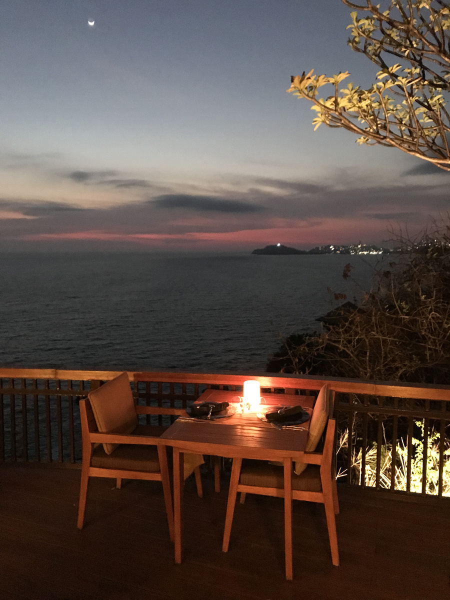 A table for two glows above the dark Pacific under a crescent moon, with Acapulco lights on the distant headland.