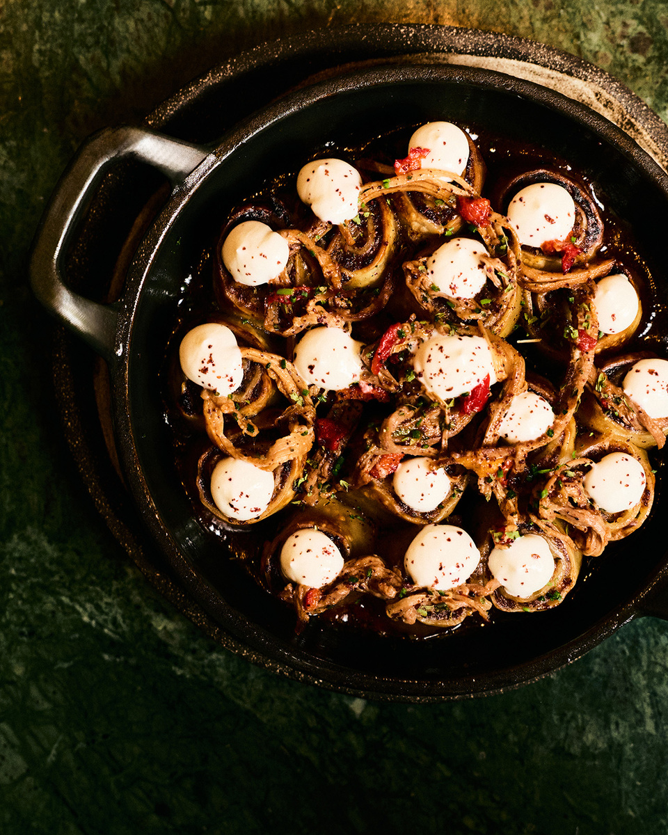 An overhead view of an Avlu dish in a dark casserole, topped with pale cream, charred onion rings, red pepper and herbs.
