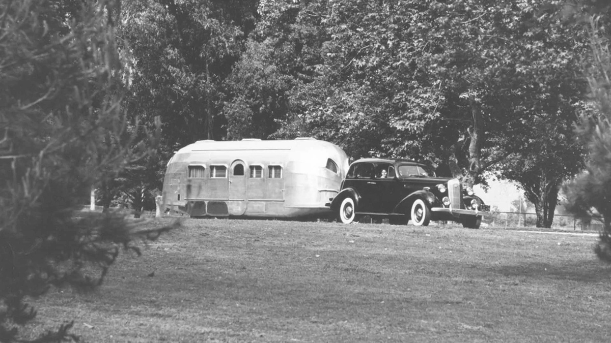 A driver sits in a dark 1937 Buick hitched to a polished aluminium Airstream Clipper on a lawn beneath trees.