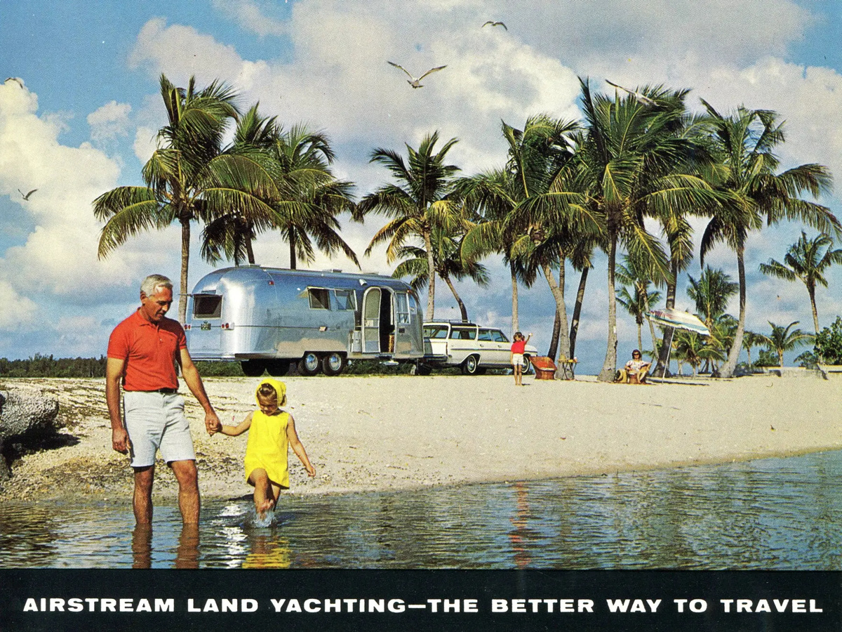 A 1960s color postcard showing a father and small daughter wading beside a tropical beach, with a silver Airstream trailer and station wagon beneath palm trees.
