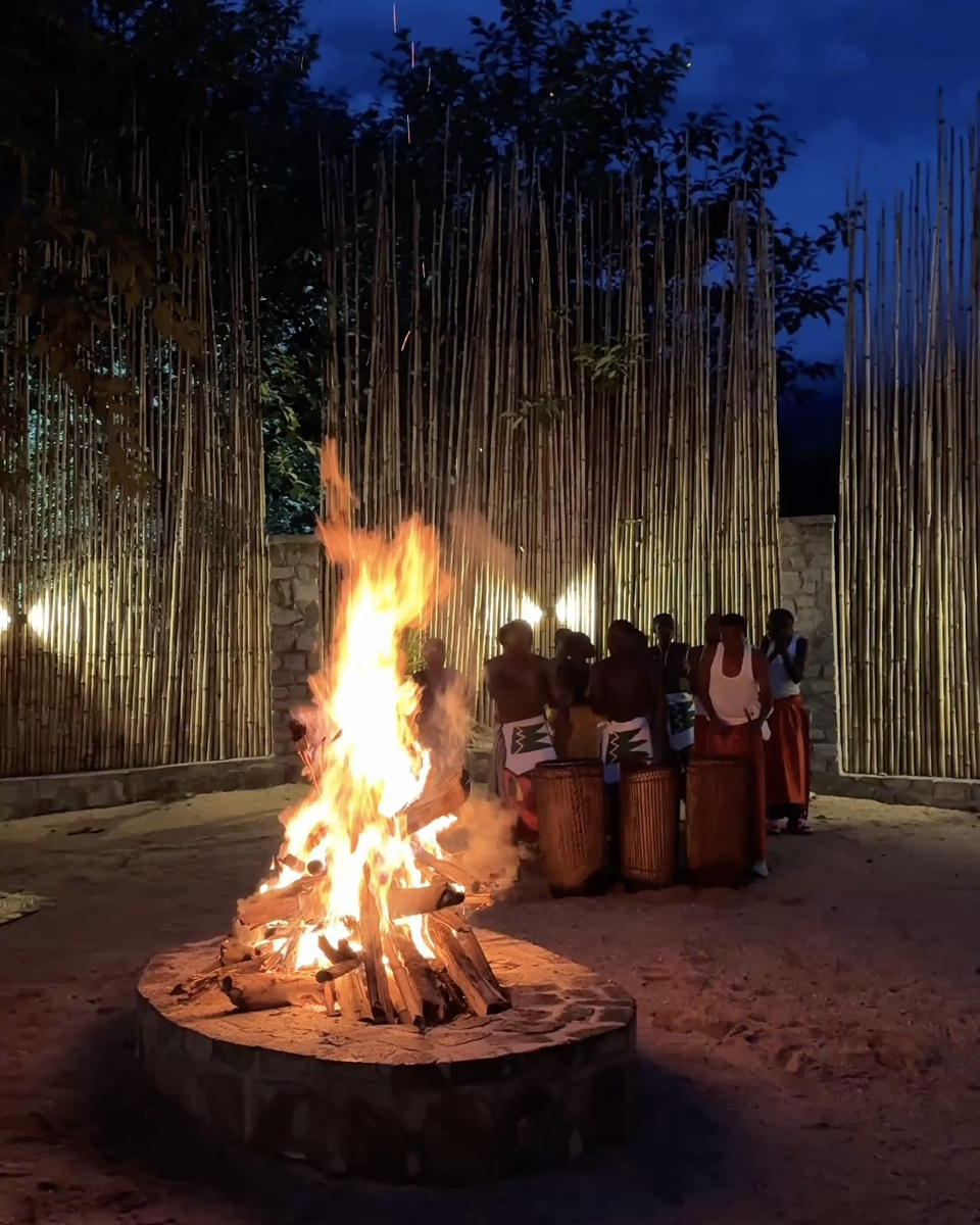 A bonfire burns in the foreground while drummers and dancers sit against a tall bamboo wall after dark.