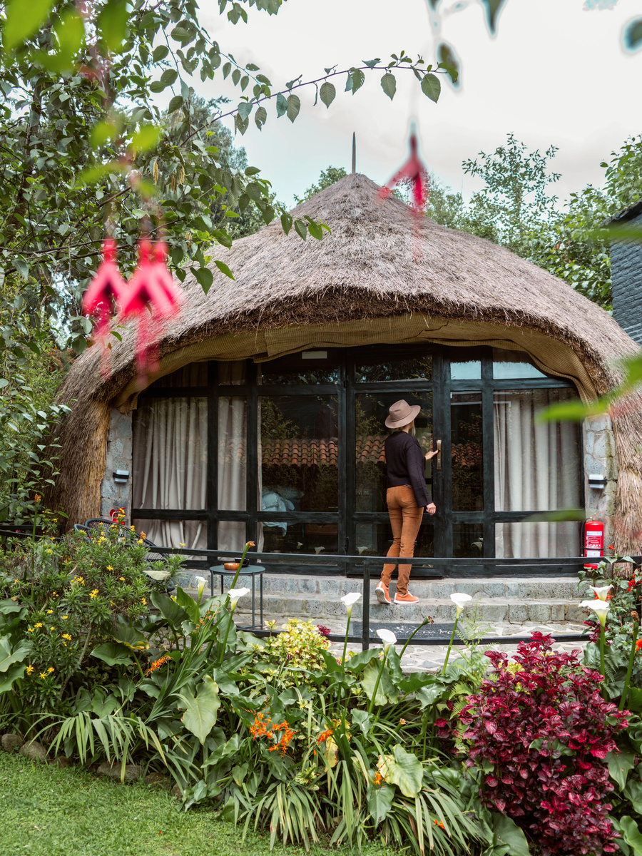 Olga stands on the terrace of a round thatched cottage behind bright garden flowers.