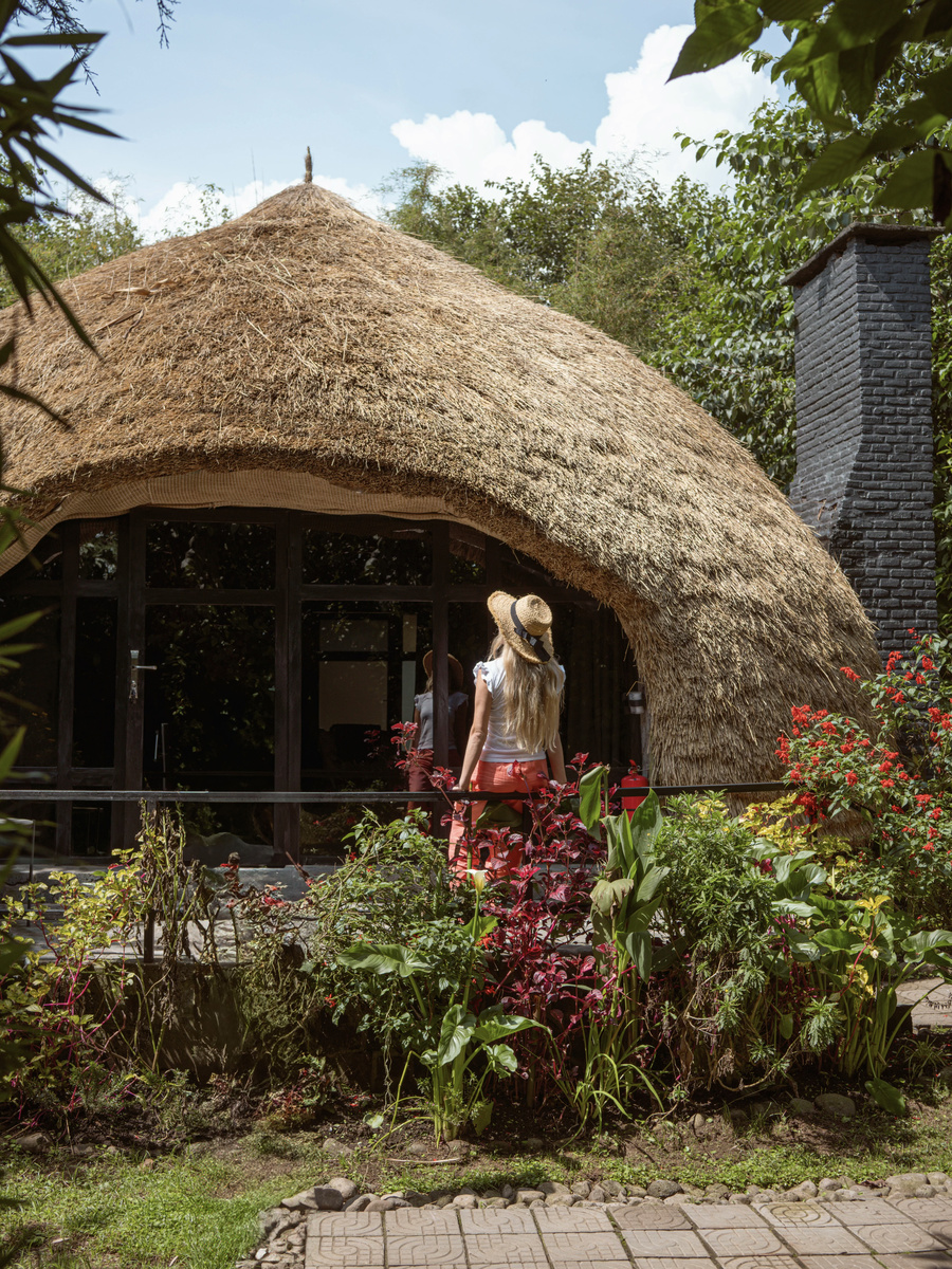 Olga stands at the open door of a thatched Amarembo cottage framed by red and orange flowers.