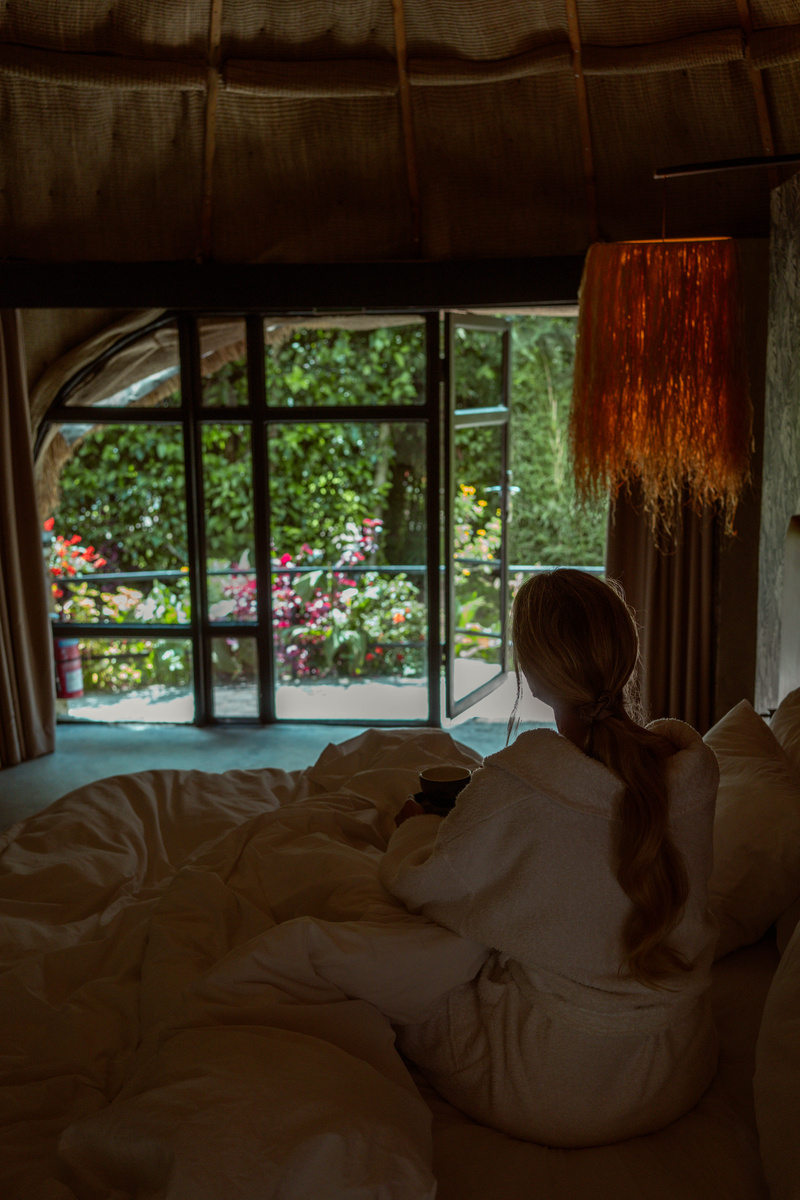 Olga sits beneath white bed linen in a thatched Amarembo cottage with the flower garden visible through the window.