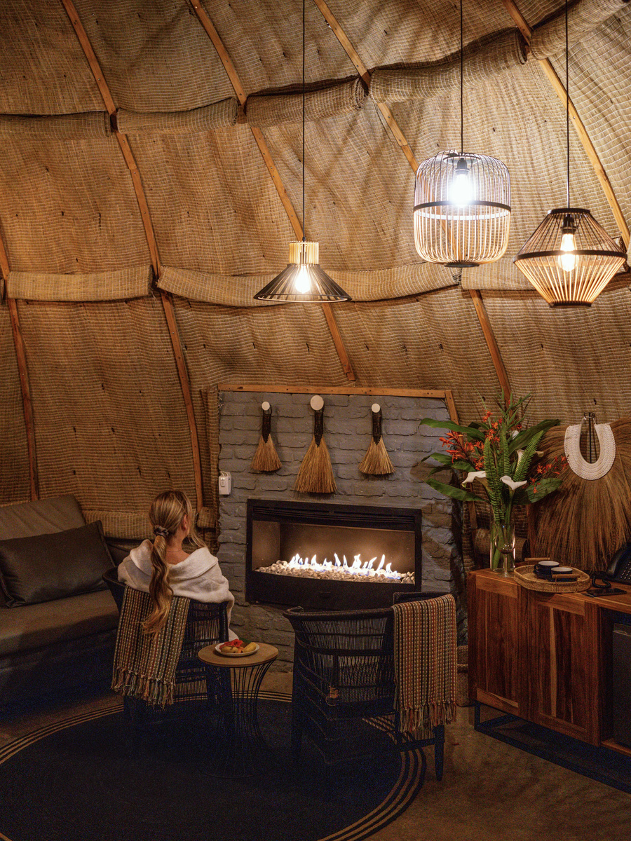 Olga sits beside a fireplace in a round room lined with woven reeds and pendant lights.
