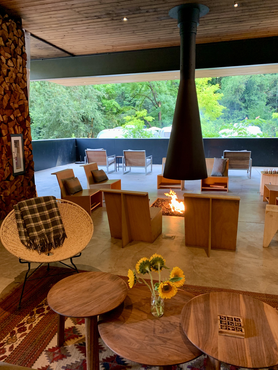 Mid-century-style clubhouse lounge with a suspended fireplace and redwood forest beyond the windows