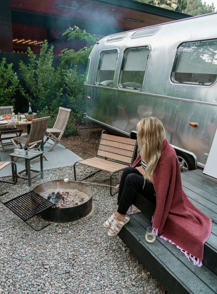 Olga sitting beside a smoking firepit outside a silver Airstream trailer at dusk