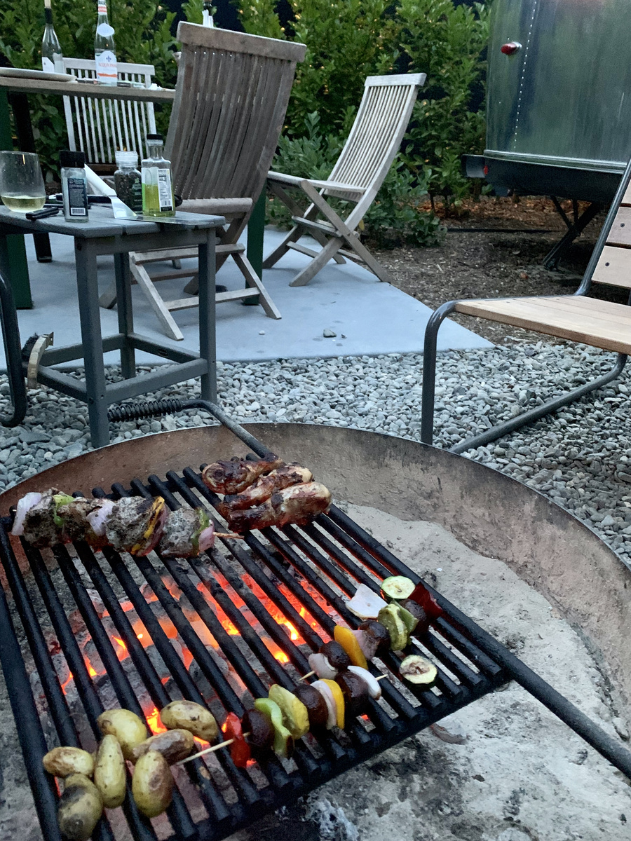 Vegetable skewers cooking over a round firepit grill outside an Airstream trailer