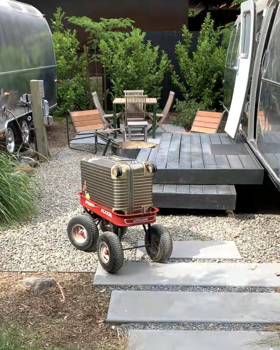Red wagon carrying a silver RIMOWA trunk along a gravel path between Airstream trailers