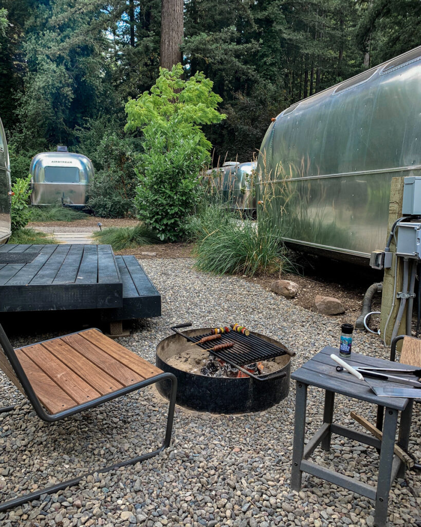 Silver Airstream trailer beside a private firepit and wooden chairs among redwoods at AutoCamp Russian River