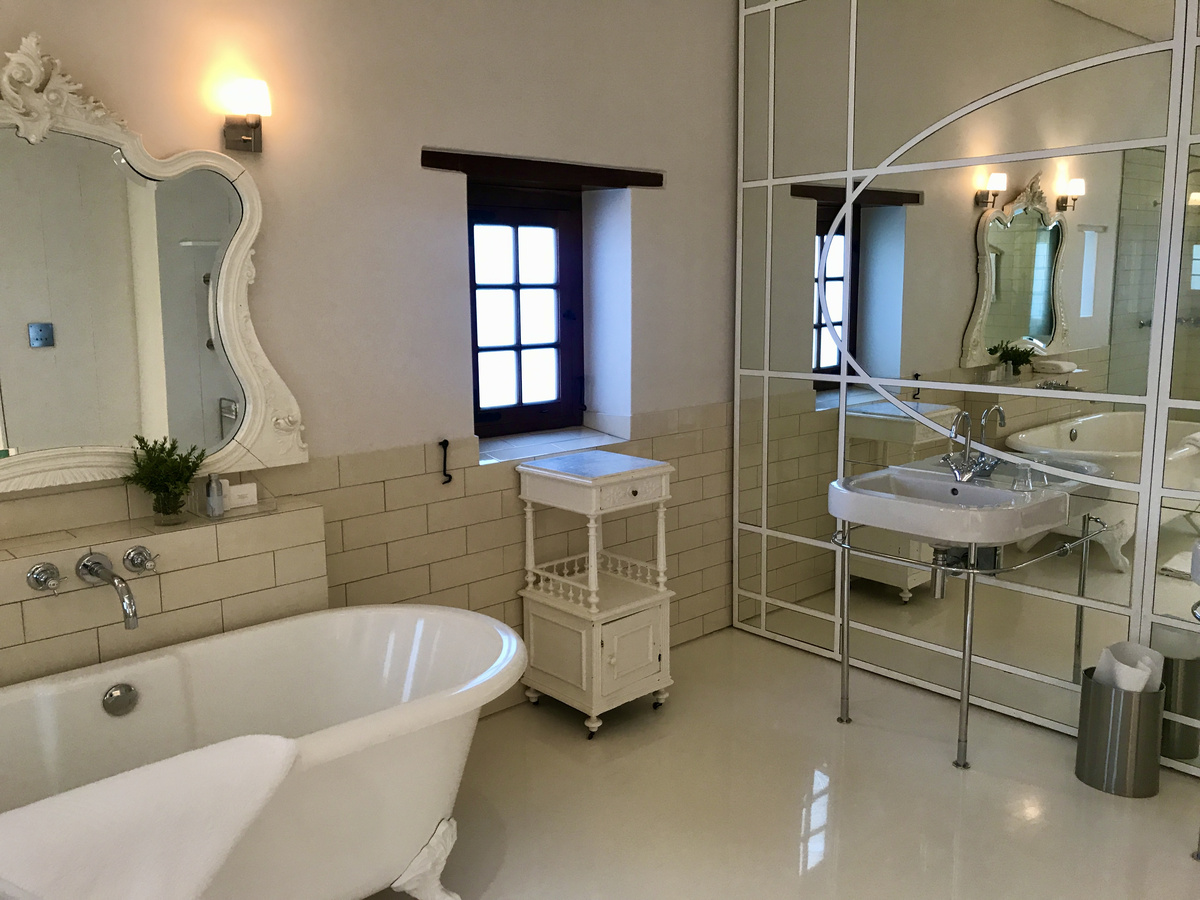 A white freestanding bath, dark-framed window and geometric mirrored wall in a Babylonstoren cottage bathroom.