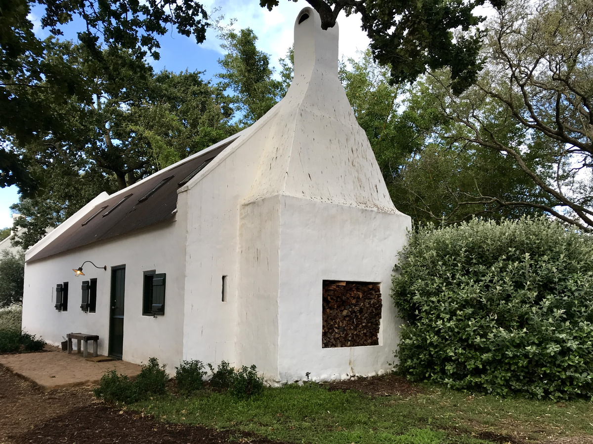 A whitewashed Babylonstoren cottage with dark shutters, a curved chimney and firewood stacked beneath oak trees.