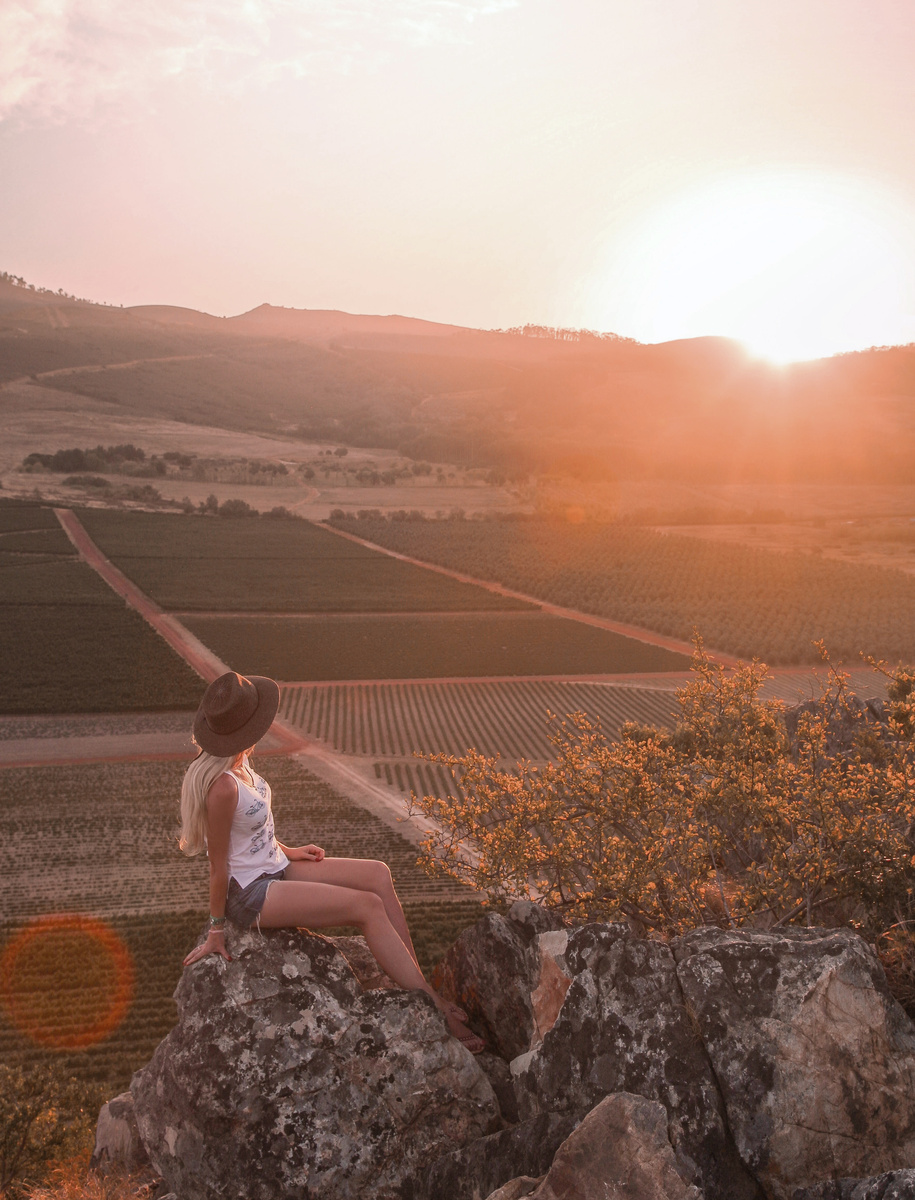 Olga sits on a rock on Koppie as the sun sets over vineyard rows below.