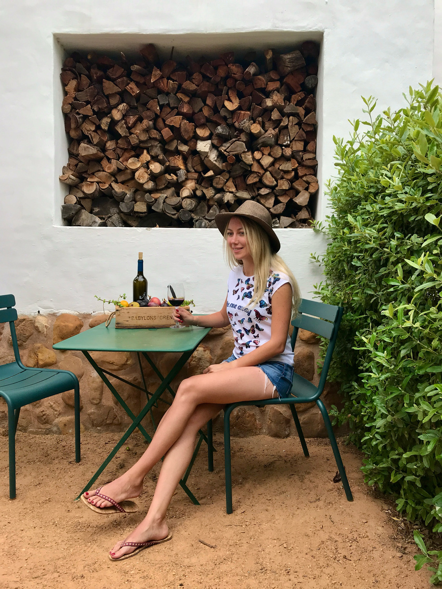 Olga sits with wine and a fruit crate at a green table beside a cottage firewood niche.