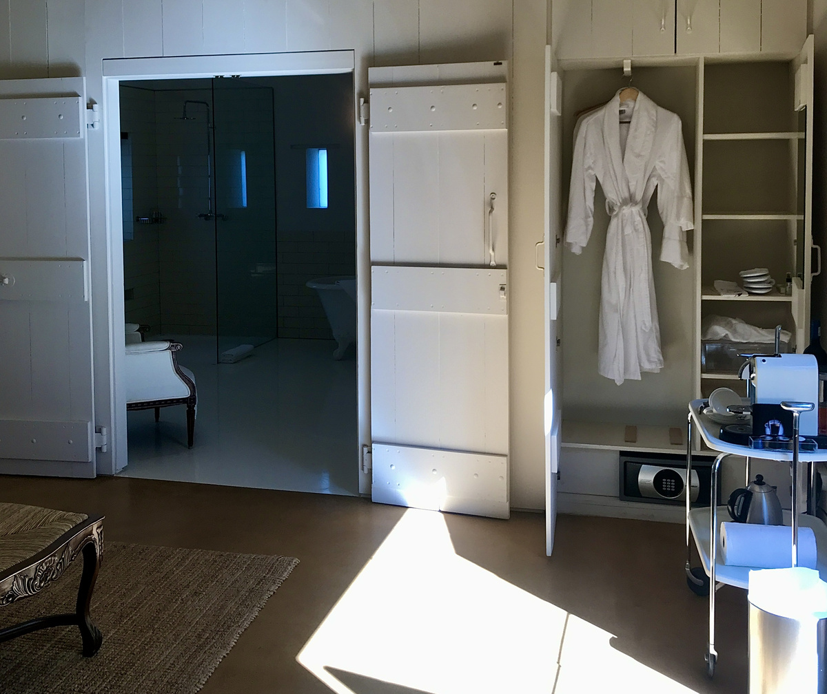 Open white cupboards hold a robe and coffee tray beside the doorway to a Babylonstoren cottage bathroom.